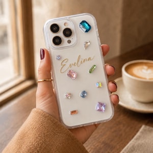 May include: A clear phone case featuring the name Evelina in gold script, decorated with various colorful, multi-shaped rhinestones. The text Made to Be Yours is visible at the top. The case has a cutout for a triple-lens camera system.