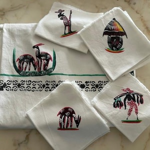 Peut inclure: Un ensemble de tissus blancs comprenant une grande nappe rectangulaire et quatre serviettes carrées. Chaque pièce est ornée d'illustrations peintes à la main en noir, rouge et vert représentant des personnages et des animaux.