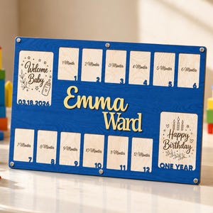 Könnte beinhalten: Ein rechteckiges blaues Holzbrett für Baby-Meilensteinfotos mit dem Namen Emma Ward in goldener Schrift. Es enthält zwölf Fächer von 1 Month bis 12 Months, einen Welcome Baby-Bereich mit dem Datum 03.18.2026 und einen Happy Birthday One Year-Bereich.