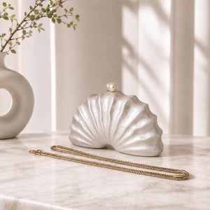 Peut inclure: Une pochette semi-circulaire avec une finition blanche nacrée et une texture côtelée en forme de coquillage. Le sac présente un fermoir perlé rond unique au centre supérieur et comprend une fine bandoulière en chaîne métallique dorée.