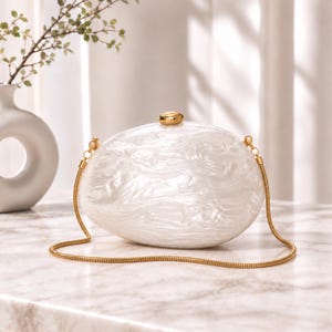 May include: An oval-shaped clutch bag with a white pearlescent marbled finish. The bag features a gold-toned metal clasp on top and a thin, flexible gold-toned snake chain shoulder strap attached to both sides of the hard-shell exterior.