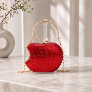 May include: A red apple-shaped clutch bag covered in small sparkling crystals. The bag features a gold-tone metal handle and a matching gold-tone chain shoulder strap. A small gold-tone stem with a clear crystal accent sits on the top centre.