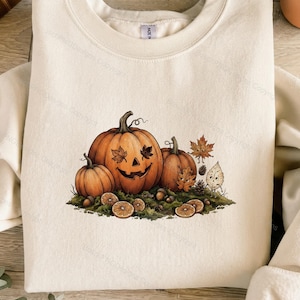 Puede incluir: Una sudadera de cuello redondo color crema con un gráfico central de tres calabazas naranjas, una tallada como linterna con hojas por ojos. El diseño incluye pequeñas hojas antropomórficas, bellotas y rodajas de naranja sobre musgo.