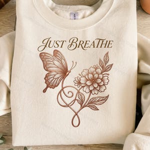 Puede incluir: Una sudadera de cuello redondo color crema con un gráfico de líneas marrones de una mariposa y una flor conectadas por un tallo en espiral. El texto "JUST BREATHE" está impreso en una fuente serif marrón sobre la ilustración.