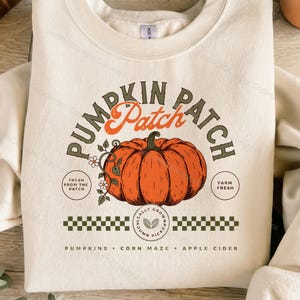 Puede incluir: Una sudadera de cuello redondo color crema con un gráfico grande de una calabaza naranja en el centro. El texto dice PUMPKIN PATCH, FRESH FROM THE PATCH, FARM FRESH y PUMPKINS, CORN MAZE, APPLE CIDER, con un patrón de cuadros y un icono de hoja.