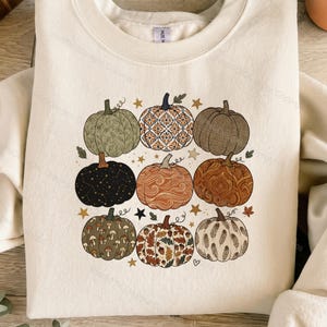 Puede incluir: Una sudadera de cuello redondo color crema con un gráfico central de nueve calabazas dispuestas en cuadrícula. Cada calabaza muestra un patrón único, incluyendo diseños florales, geométricos, celestiales, de setas, hojas y plumas con estrellas.