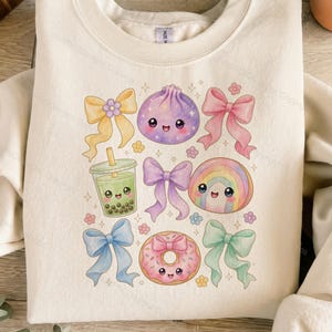Puede incluir: Una sudadera de cuello redondo color crema con un estampado gráfico central de varios elementos estilo kawaii, incluyendo una bola de masa morada, un té de burbujas verde, un arcoíris y una dona rosa, rodeados de lazos y flores pequeñas.