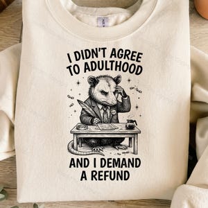 Puede incluir: Una sudadera de cuello redondo color crema con un gráfico negro de una zarigüeya con traje, sentada en un escritorio con una pluma y café. El texto dice I DIDN'T AGREE TO ADULTHOOD AND I DEMAND A REFUND en una fuente negra y negrita.