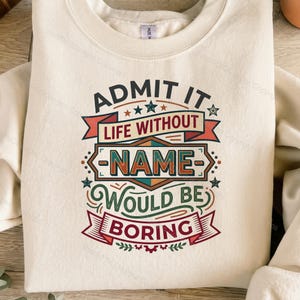 Puede incluir: Una sudadera de cuello redondo color crema con un diseño gráfico que incluye el texto "ADMIT IT LIFE WITHOUT NAME WOULD BE BORING" en azul oscuro, rojo y verde. El diseño presenta pancartas decorativas, estrellas y formas.