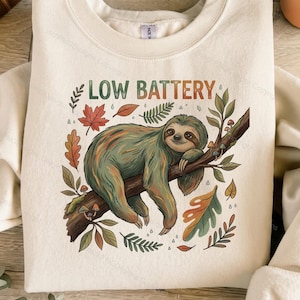 Op de afbeelding: Een crèmekleurig sweatshirt met ronde hals en een afbeelding van een luiaard die op een boomtak rust. De tekst LOW BATTERY staat erboven. Het ontwerp bevat kleurrijke herfstbladeren, kleine paddenstoelen en regendruppels in aardetinten.