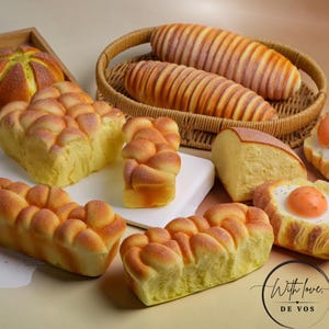 May include: A collection of various golden-brown baked bread items, including braided loaves, a round bun, and a pastry topped with a fried egg, arranged with two long loaves in a woven basket and the text With love DE VOS in the corner.