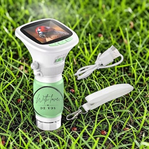 May include: A white digital microscope with a screen displaying a ladybug, featuring a light green textured grip printed with the text With love, DE VOS. The device includes a white USB charging cable and a white plastic clip attachment.
