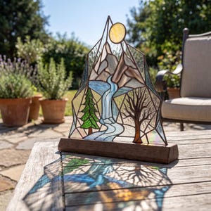 May include: A triangular stained glass panel featuring a mountain landscape with a waterfall, a green pine tree, a bare brown tree, and a yellow sun, all set within a dark metal frame and mounted on a rectangular wooden base.