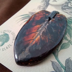 May include: A glossy, leaf-shaped pendant featuring a dark brown and amber glaze with visible veining. The pendant has a smooth, reflective surface and a small hole at the top for a cord. It rests on a card with the text LIMITED EDITION GLAZE.