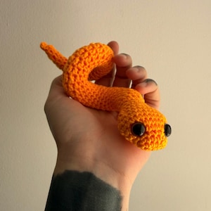 Può includere: Un serpente giocattolo all'uncinetto di colore arancione brillante con una superficie in filato testurizzato e due grandi occhi rotondi in plastica nera lucida. Il serpente è avvolto in una forma curva, mostrando l'intricato motivo a maglia.