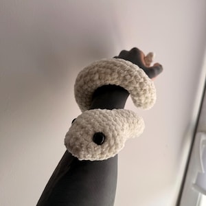 May include: A cream-coloured crocheted snake plush toy with a soft, textured yarn finish. The snake features two small, round, black plastic eyes and a long, coiled body that wraps around an arm, showcasing its flexible and plush construction.