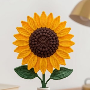 Puede incluir: Un girasol de ganchillo con pétalos de color amarillo brillante y un centro marrón oscuro, que presenta dos hojas verdes en un tallo. La flor se exhibe en un pequeño jarrón de cerámica blanca lisa, creando una pieza decorativa de colores vivos.