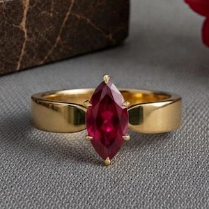 May include: A gold ring featuring a marquise-cut deep red gemstone set in the centre. The band is smooth and polished, with a solid gold finish that tapers slightly towards the setting. The gemstone is held in place by small metal prongs.