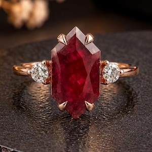 May include: A rose gold ring featuring a large, hexagonal-cut deep red gemstone as the center stone, flanked by two small, round, clear white accent stones. The ring is set in a six-prong setting for the center stone and a four-prong setting for each side.