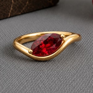 Peut inclure: Une bague en or ornée d'une pierre précieuse rouge foncé taillée en marquise, montée sur un anneau lisse et incurvé. Le métal présente une finition polie et la pierre est placée au centre de la monture, créant un design fluide et élégant.