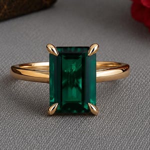 May include: A gold ring featuring a rectangular emerald-cut green gemstone set in a four-prong setting. The band is polished gold, and the stone is a deep, vibrant green color, positioned centrally to showcase the clarity and cut of the gemstone.