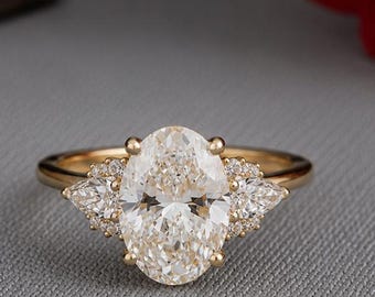 Pode incluir: Um anel de ouro com uma grande pedra central de corte oval, ladeada por duas pedras menores de corte marquise em cada lado. A cravação inclui pequenas pedras redondas ao redor da base do oval central, criando um design detalhado.