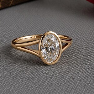 May include: A gold ring featuring a central oval-cut clear gemstone set in a smooth bezel setting. The band is crafted with a split-shank design that tapers towards the centre stone, creating a polished and refined look with a warm metallic finish.