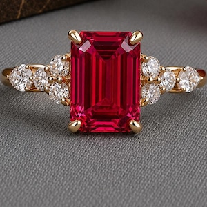 Puede incluir: Un anillo de oro con una gran piedra preciosa roja de corte esmeralda rectangular en el centro, flanqueada por tres pequeños diamantes blancos redondos a cada lado. La banda está hecha de oro amarillo pulido con un engaste clásico.
