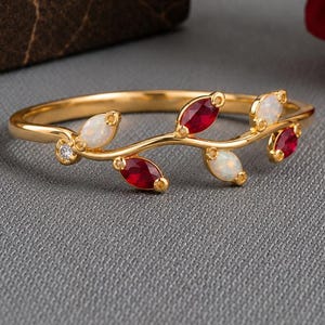 May include: A gold ring featuring a delicate vine-like band adorned with alternating marquise-cut red rubies and white opals, accented by a single small round clear gemstone at the base of the leaf-shaped arrangement.