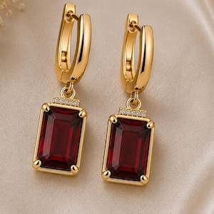 May include: A pair of gold hoop earrings featuring rectangular, deep red faceted gemstones suspended from each hoop. Each earring includes a small, horizontal bar set with tiny clear stones positioned directly above the large red gemstone.