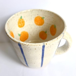 Op de afbeelding: Een keramische mok met een gespikkeld crèmekleurig glazuur, voorzien van handgeschilderde oranje fruitmotieven aan de binnenzijde en verticale blauwe strepen aan de buitenzijde. De mok heeft een brede, ronde komvorm en een stevig handvat.