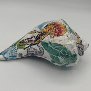 May include: A white conch shell decorated with colorful illustrations of marine life, including a yellow and orange jellyfish, a green sea turtle, a blue seahorse, and various seashells, all rendered in a detailed, artistic style across the surface.