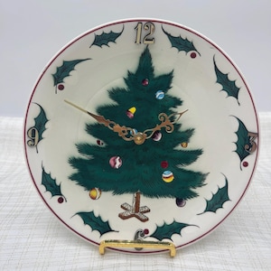 May include: A round ceramic wall clock with a white background featuring a central green Christmas tree decorated with colorful ornaments. The clock face displays numbers 3, 6, 9, and 12, accented by green holly leaves and red berries.