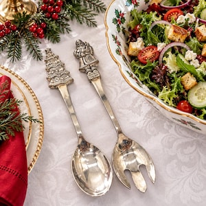 May include: A pair of silver-toned metal salad servers featuring decorative handles. One handle is shaped like a Christmas tree, and the other is shaped like Santa Claus. The set includes a solid spoon and a three-pronged fork for serving salad.