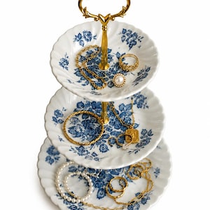 May include: A three-tiered white porcelain serving stand with a scalloped edge and blue floral pattern, featuring a gold-tone metal central handle. The tiers are filled with various gold-tone necklaces, rings, and a strand of white pearls.
