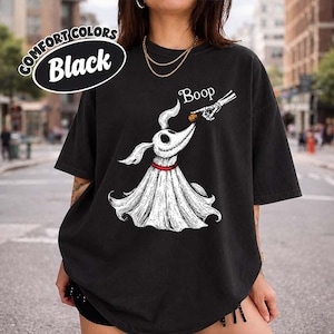 May include: A black t-shirt featuring a white graphic of a ghost dog with a red collar, a skeleton hand touching its nose, and the text Boop. An oval label in the top left corner reads Comfort Colors Black. The shirt has short sleeves.
