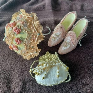 May include: A miniature decorative set featuring a pale yellow parasol with lace trim and pink floral accents, a pair of light pink lace-covered slippers with pearl embellishments, and a small white fabric purse with gold and pearl bead trim.
