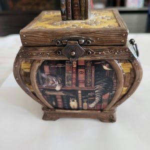 Peut inclure: Une boîte décorative imitation bois avec un loquet métallique articulé. Le couvercle présente une sculpture miniature de livres. Le panneau avant affiche une illustration imprimée d'un chat dormant sur une étagère remplie de livres et d'un hibou.