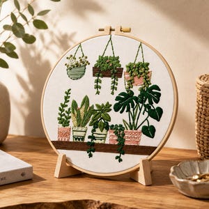 Peut inclure: Un tambour à broder circulaire en bois contenant un tissu blanc avec un motif cousu à la main représentant diverses plantes en pot. La broderie présente trois jardinières suspendues et cinq plantes sur une étagère, dans des tons de vert, rose et marron.