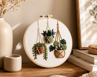 May include: A circular wooden embroidery hoop containing white fabric with a hand-stitched design of three hanging potted plants. The plants are rendered in shades of green, brown, and tan thread, featuring various leaf shapes and macrame-style hangers.