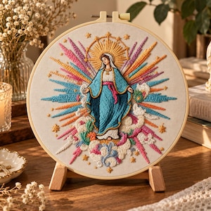 Peut inclure: Un tambour à broder circulaire présentant une représentation cousue à la main de la Vierge Marie en robe bleue et blanche, entourée d'un rayon de soleil multicolore vibrant, de petites étoiles jaunes et de nuages blancs sur tissu crème.