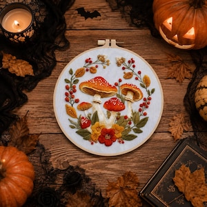 Peut inclure: Un tambour à broder circulaire en bois contenant un panneau en tissu blanc avec un motif brodé à la main de champignons rouges et orange, de feuilles vertes, de fleurs jaunes et de petites baies rouges disposées en couronne.