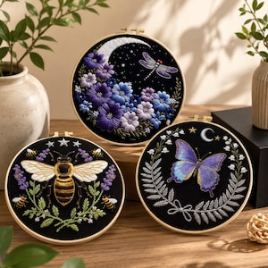 Könnte beinhalten: Drei runde Stickrahmen aus Holz mit schwarzem Stoffhintergrund. Ein Rahmen zeigt eine Mondsichel mit violetten und blauen Blumen sowie eine Libelle. Ein weiterer zeigt eine große Biene mit Lavendelzweigen und Sternen. Der dritte zeigt einen violetten Schmetterling, Maiglöckchen und einen kleinen Mond.