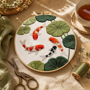 Peut inclure: Un tambour à broder circulaire en bois contenant un panneau de tissu blanc avec des carpes koï brodées à la main en orange, blanc et noir, entourées de nénuphars verts. Le motif représente trois koïs nageant parmi le feuillage.