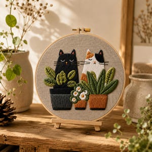 Peut inclure: Un tambour à broder circulaire représentant un chat noir et un chat calicot assis derrière des plantes en pot. Le chat noir est dans un pot gris foncé, tandis que le chat calicot est à côté d'un pot en terre cuite avec des fleurs blanches.