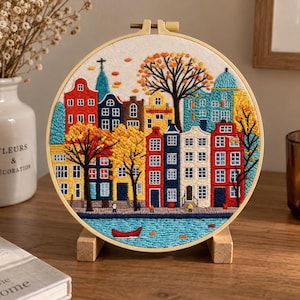 Pode incluir: Um bastidor de bordado circular com uma cena colorida costurada de casas de canal no outono. O design inclui edifícios vermelhos, amarelos, azuis e brancos, árvores com folhas laranja e amarelas e um pequeno barco vermelho num canal azul.
