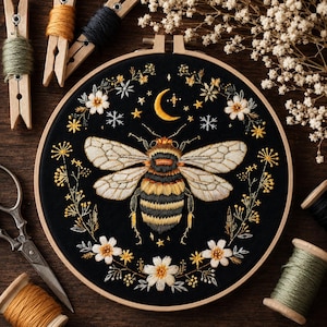 Peut inclure: Un tambour à broder circulaire en bois contenant un tissu noir avec un bourdon brodé à la main au centre. L'insecte est entouré d'une couronne de fleurs blanches, d'étoiles jaunes et d'un croissant de lune, réalisés en fil à broder.