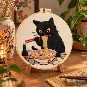 May include: A circular embroidery hoop featuring a black cat with yellow eyes eating noodles from a white bowl with red patterns using red chopsticks. The scene includes a small bottle, a green container, and side dishes on a cream fabric background.