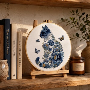 Peut inclure: Un tambour à broder circulaire en bois contenant un tissu blanc avec un motif de chat détaillé composé de diverses fleurs bleues et blanches, agrémenté de trois petits papillons dans des tons de bleu et de jaune.