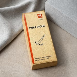 Könnte beinhalten: Ein rechteckiger hellbrauner Karton für einen Zwilling J.A. Henckels Twin Stone Schleifstein. Die Schachtel hat einen roten vertikalen Streifen auf der linken Seite, den Schriftzug TWIN STONE, eine Grafik eines Messers auf einem Stein und Text.